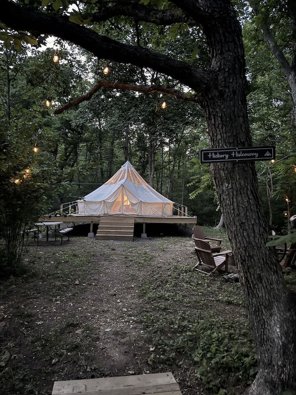 Un terrain de camping équipé d’une tente, d’une terrasse en bois et d’un espace détente, le tout entouré d’arbres et illuminé de guirlandes lumineuses.
