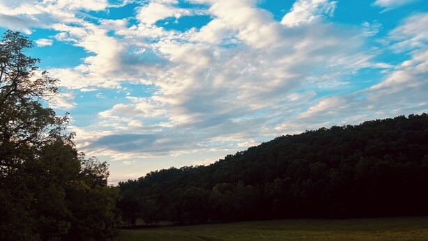Cielo despejado con algunas nubes dispersas sobre un campo cubierto de hierba y una colina boscosa.