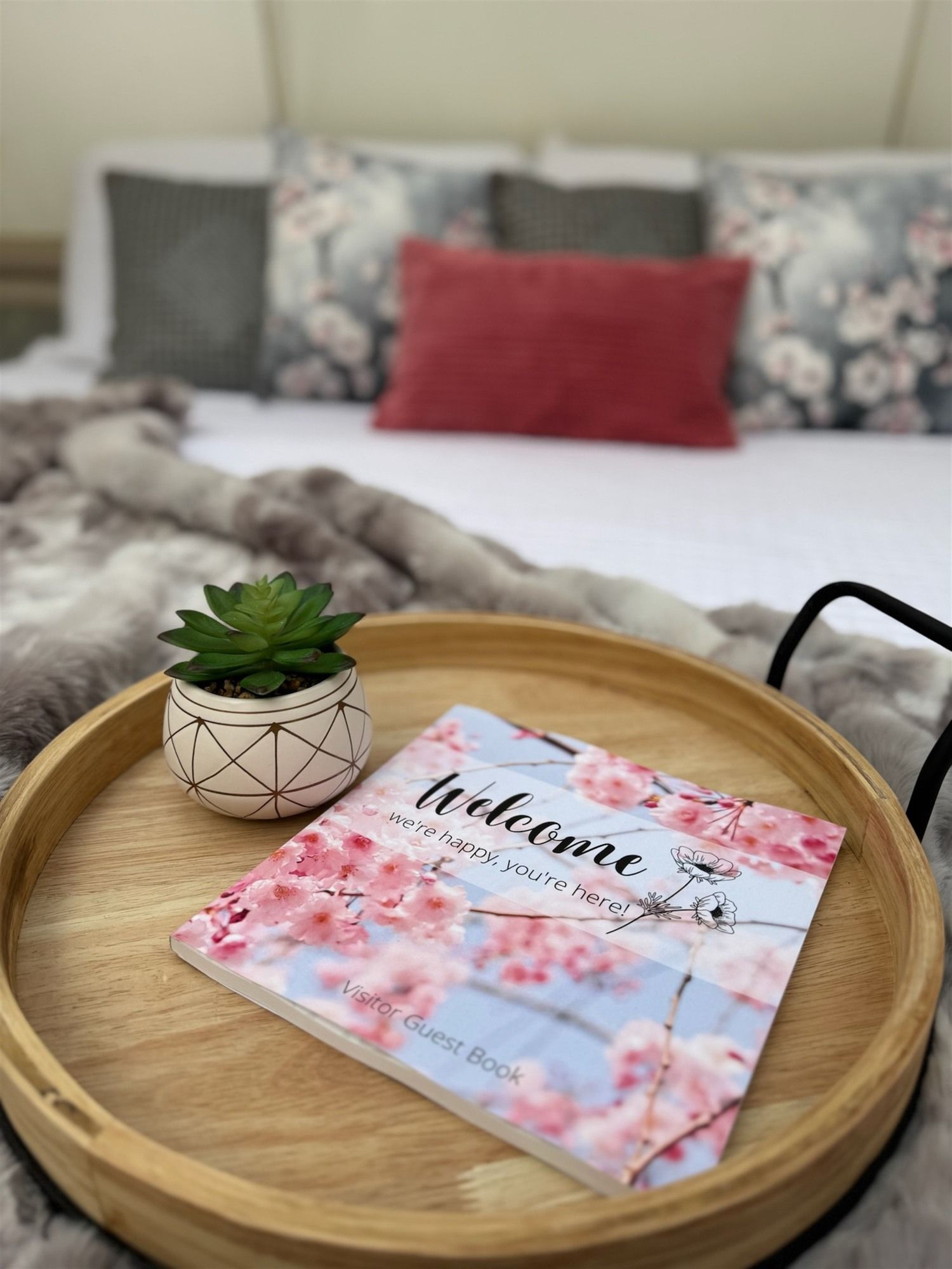 Una bandeja de madera con una tarjeta de bienvenida y una pequeña planta en maceta sobre una cama con ropa de cama estampada con motivos florales.