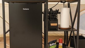 Una kitchenette compacta con un refrigerador Galanz negro, un microondas, una cafetera y un estante pequeño con condimentos y utensilios.