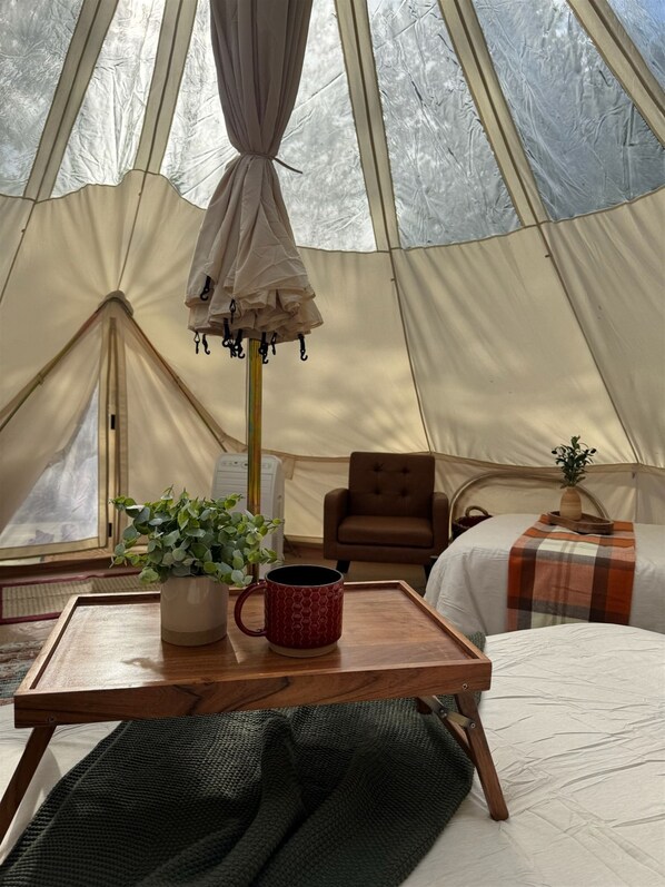 Interior de una tienda de campaña con una cama, una silla, una mesa con una taza y una planta en maceta.
