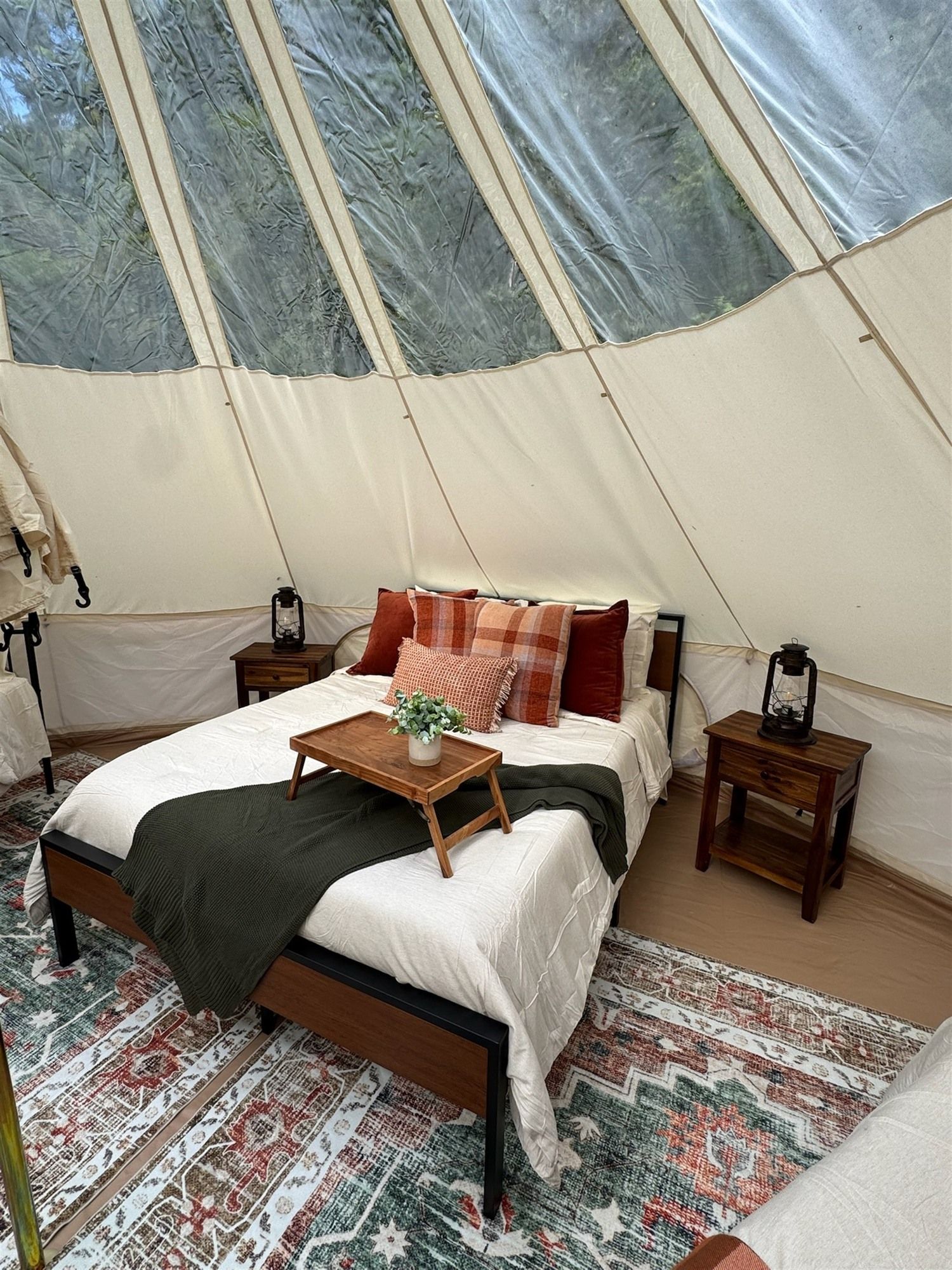 A tented accommodation with a bed, bedside tables, and a small table with a plant.