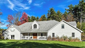 Une maison blanche à deux étages, dotée d’un porche, d’un petit jardin devant et surplombée d’un ciel bleu clair.