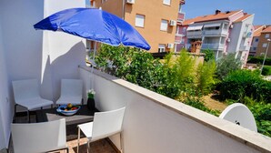 Un balcon avec des chaises blanches, une table sur laquelle se trouve un saladier rempli de fruits et un parasol bleu.