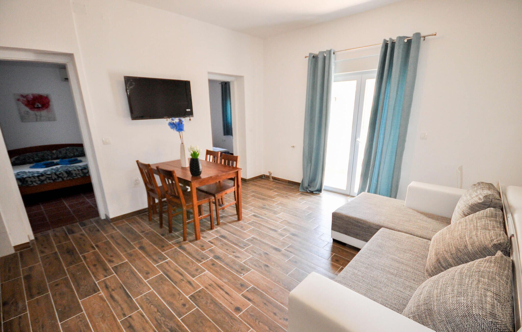 Un salon avec un plancher en bois, une table à manger avec des chaises, un canapé, un téléviseur fixé au mur et une porte-fenêtre coulissante avec des rideaux.