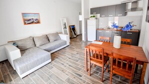 Un salon moderne avec un canapé gris, une table à manger en bois et des chaises. On aperçoit, au fond, une cuisine avec des meubles gris et un réfrigérateur blanc.
