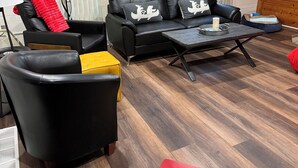 A modern living room with a black leather sofa, a yellow ottoman, and a red bench. There is a wooden coffee table with a candle and a glass. The room has wooden beams on the ceiling and large windows with blinds.