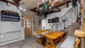 Un interior rústico con una mesa de madera y bancos, paredes de piedra, una araña y una escalera.