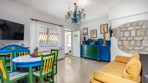Una sala de estar con un sofá, una mesa de comedor con sillas, un televisor y una zona de cocina con un mueble azul y una pared de piedra.
