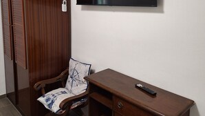 Une pièce comprenant un bureau en bois, une chaise avec un coussin, un téléviseur fixé au mur et une armoire.