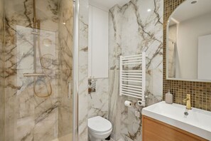 Une salle de bain avec un mur en marbre, des toilettes blanches, une douche avec une porte en verre et un lavabo blanc avec un robinet doré.