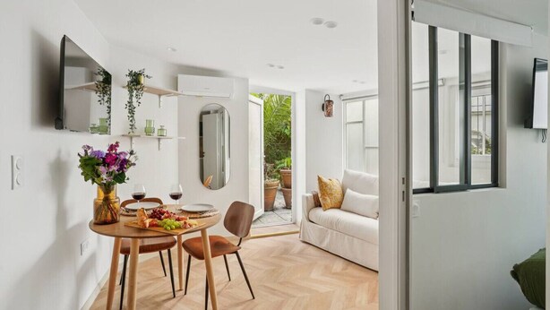 A modern kitchen with a dining area, a TV mounted on the wall, and a view of the garden through an open door. - Lavie Maison AC & Terrace (Paris)