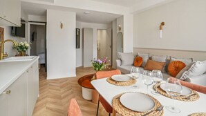 Un comedor moderno con una mesa blanca para cuatro personas, rodeado por un sofá gris con cojines naranjas.