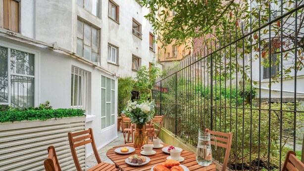 Een houten tafel gedekt met borden eten, een bloevas en een waterkan op een balkon met een metalen hek.