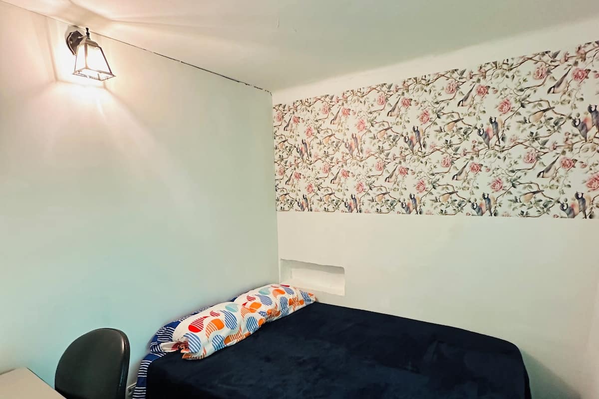 Ein kleines Zimmer mit einem Einzelbett, einem Schreibtisch, einem Stuhl, einer Wand mit Muster und einer Deckenleuchte.