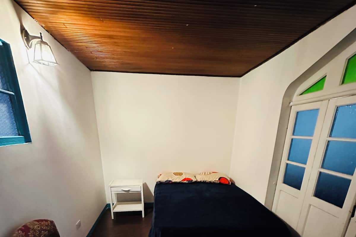 Ein kleiner, schummrig beleuchteter Raum mit einem Einzelbett, einem Nachttisch und einem Stuhl.