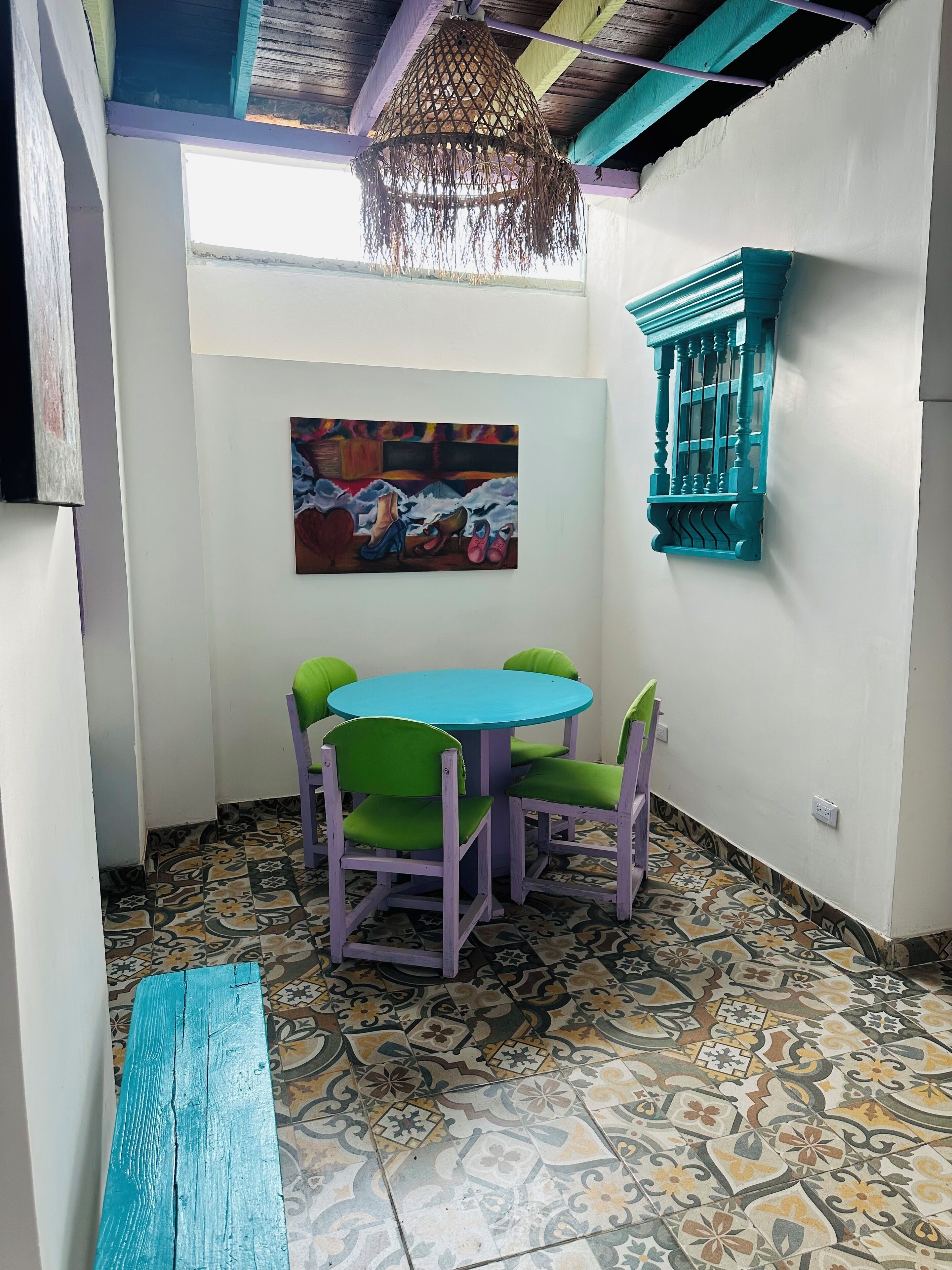 A room with a round table and four chairs, a hanging light fixture, a colorful painting on the wall, and a window with a blue frame.