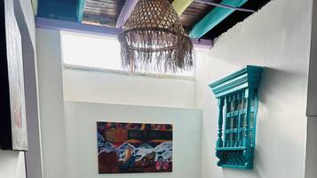 A room with a round table and four chairs, a hanging light fixture, a colorful painting on the wall, and a window with a blue frame.
