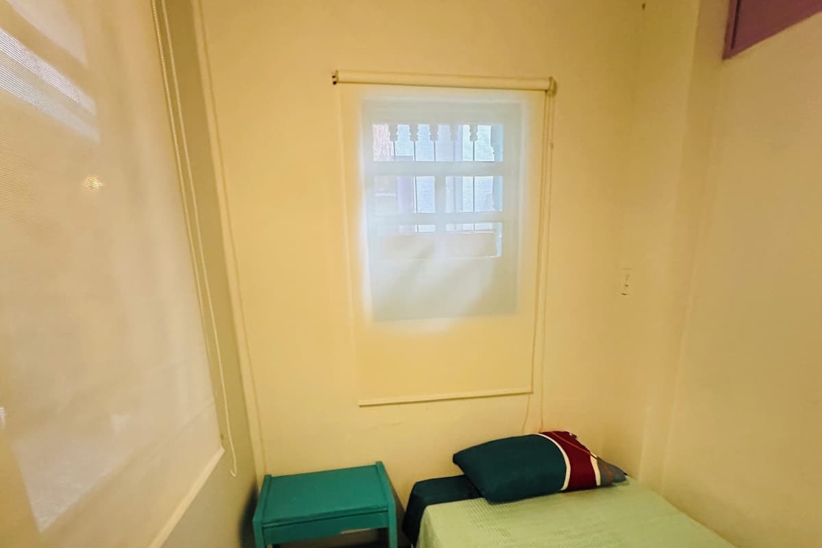Ein kleiner, schmaler Raum mit einem Einzelbett, einem Tisch und einem Fenster mit Projektionsschirm.