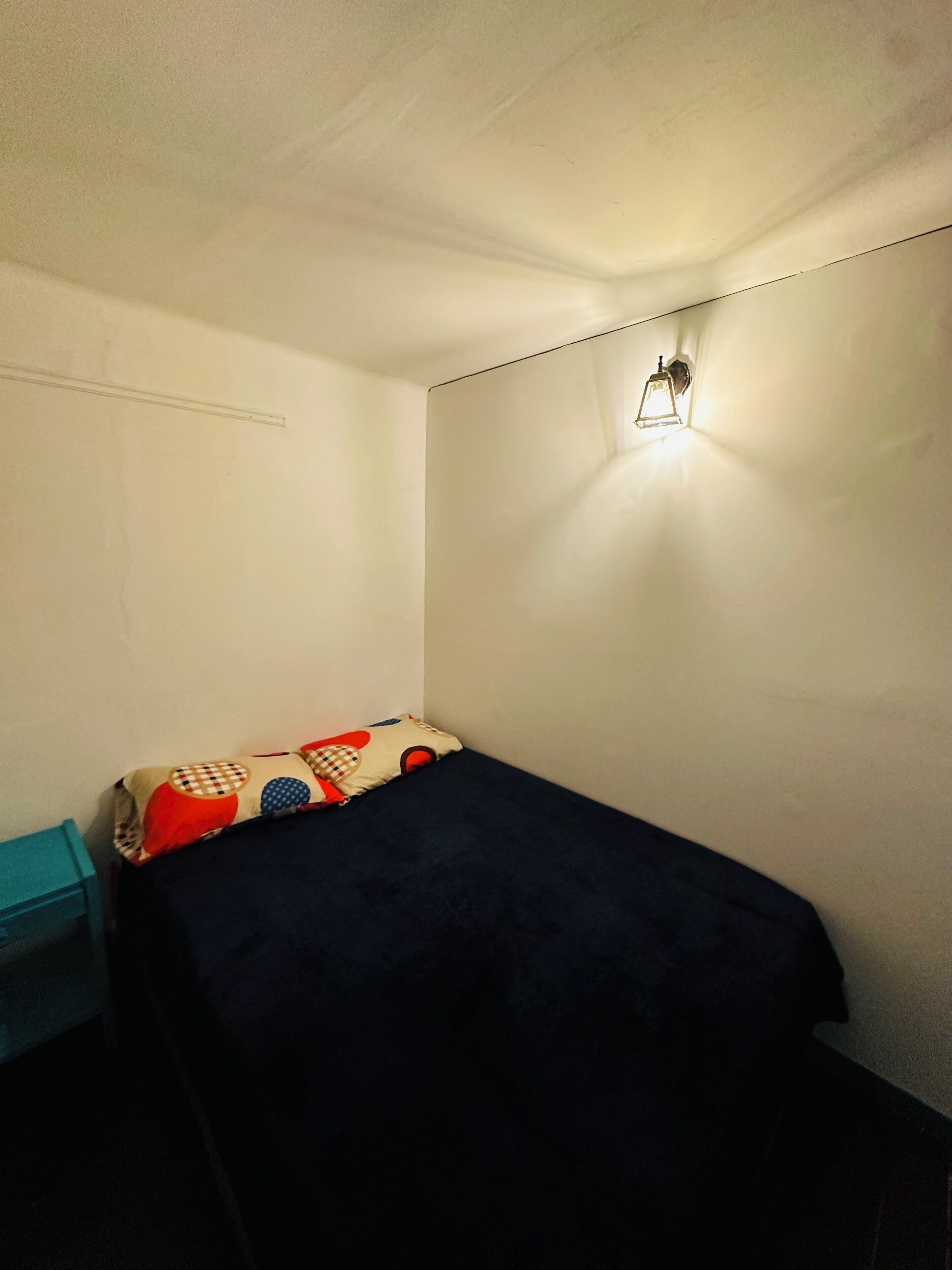 Una habitación pequeña con una cama individual, techo blanco liso, lámpara de pared y un cojín colorido en la cama.