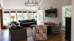 Een ruime woonkamer met een grote bank, voetenbankje en gestreept fauteuils. Een kroonluchter hangt aan het plafond, en een flatscreen televisie is aan de muur bevestigd.