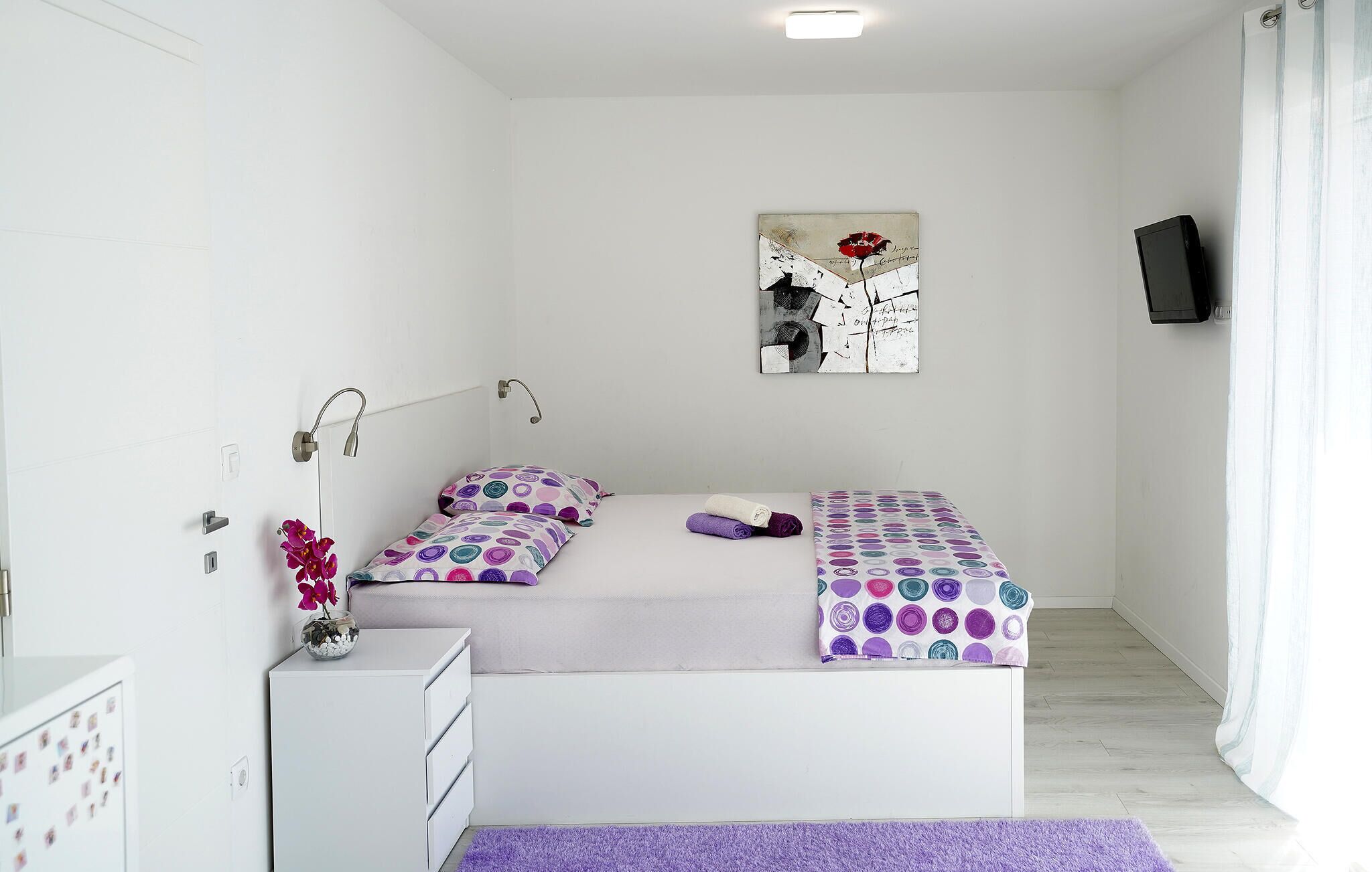 A bedroom with a bed, a bedside table, a wall-mounted TV, and a painting.