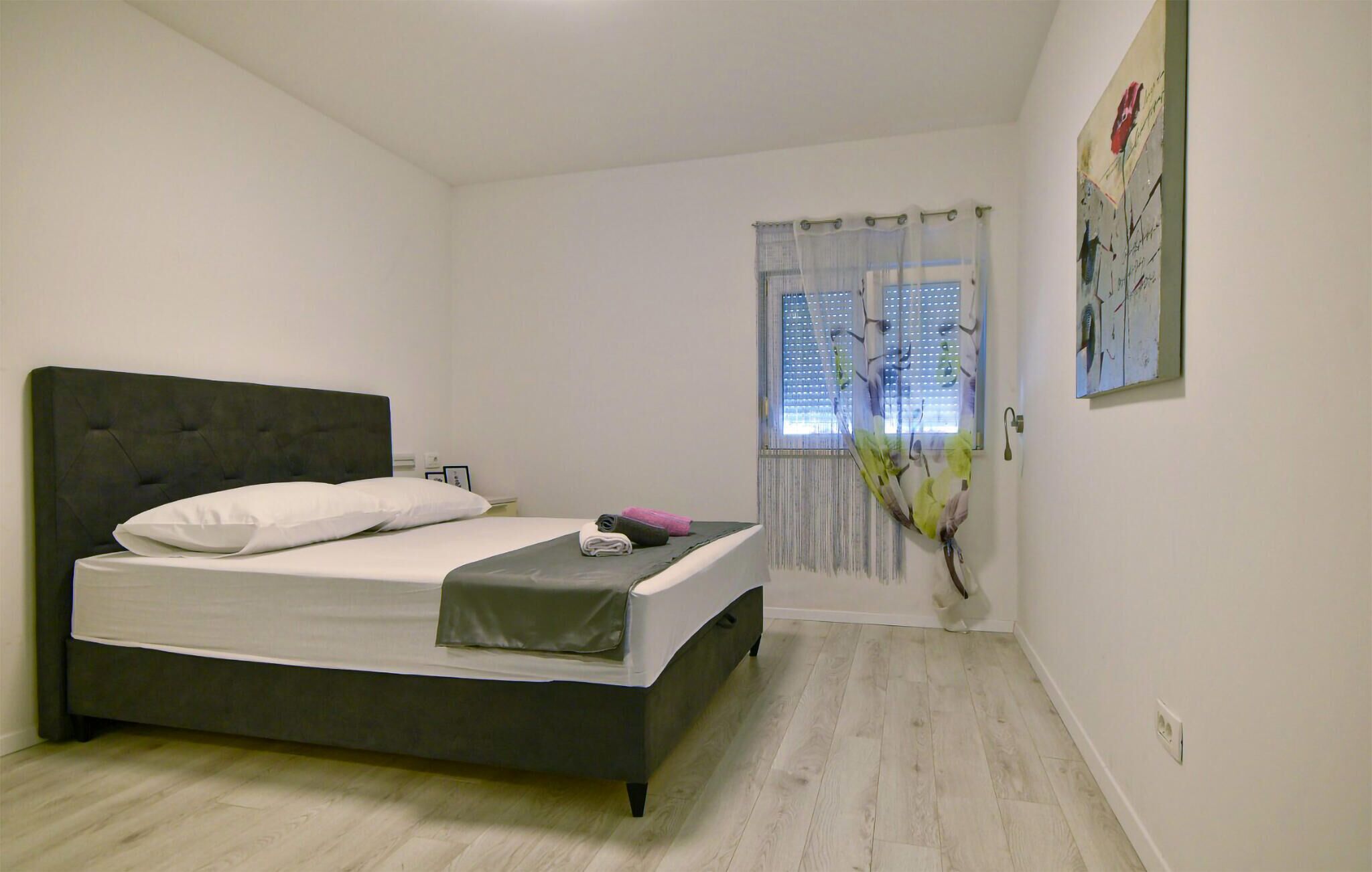 A bedroom with a bed, a nightstand, a window with curtains, and a wall-mounted artwork.