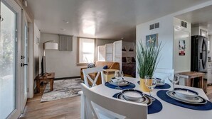 A modern dining area with a white table set for four, a sofa, and a TV in the background. - Dock&Volleyball! Near Downtown- Orange Oasis (Savannah)