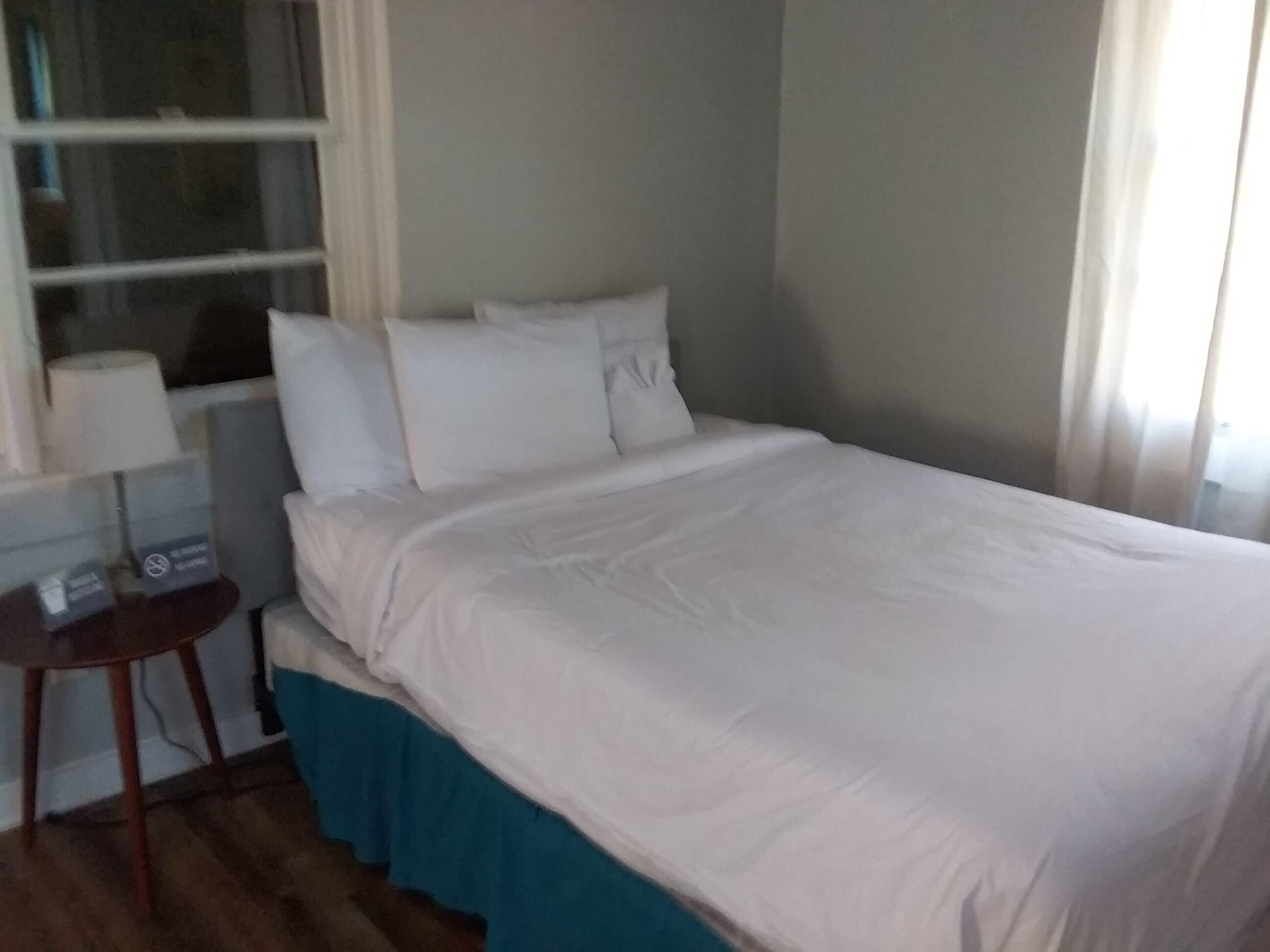 A neatly made bed with white linens in a room with a window, a small table, and a blue skirt on the bed.