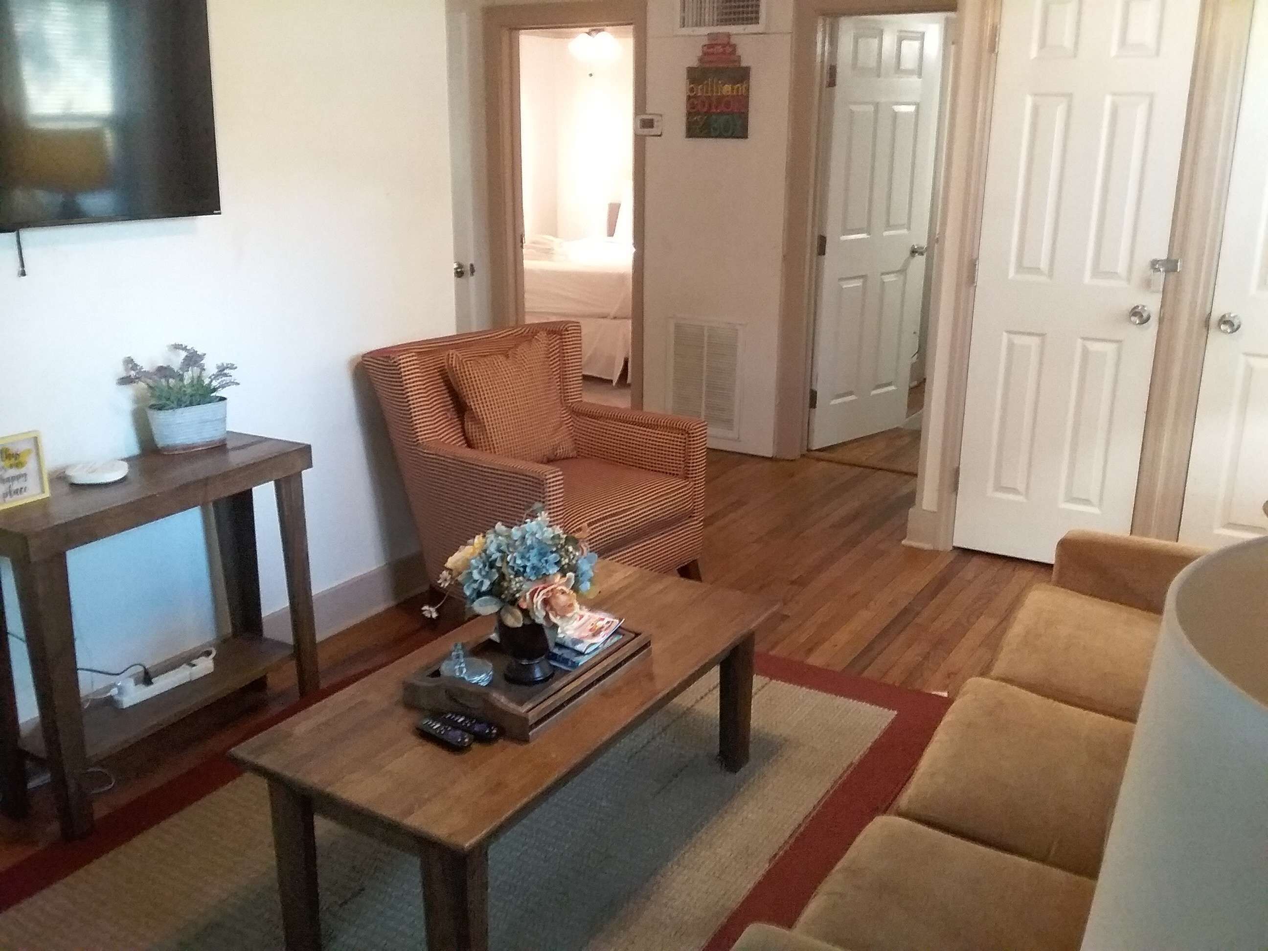 A living room with a brown sofa, a wooden coffee table with a vase and remote controls, a wicker armchair, and a television on the wall.