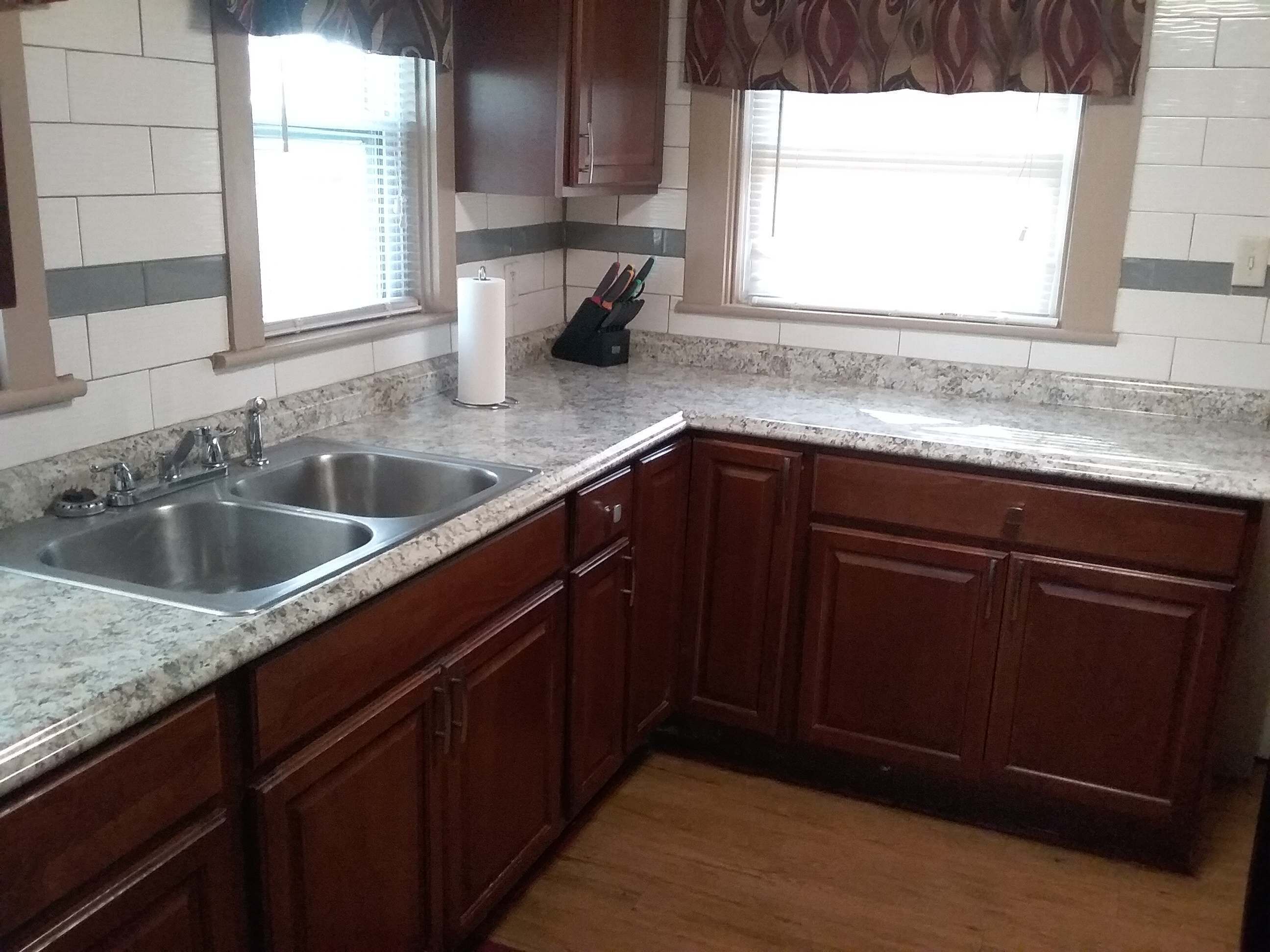 A kitchen with two sinks, wooden cabinets, a marble countertop, and a window with blinds.
