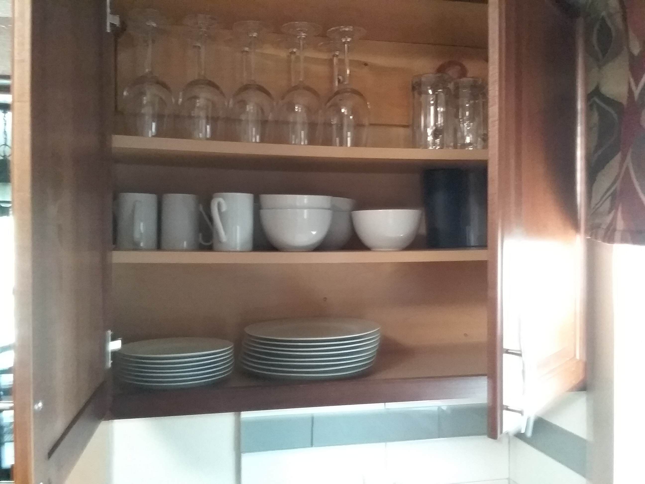 A kitchen cabinet with shelves holding glassware, white mugs, bowls, and stacked plates.