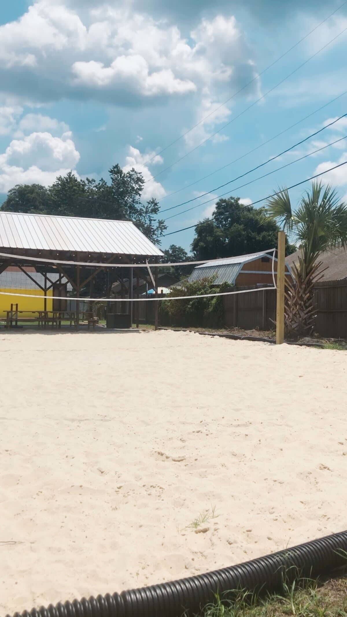 A sandy outdoor volleyball court with a metal roof structure, picnic tables, and a palm tree.