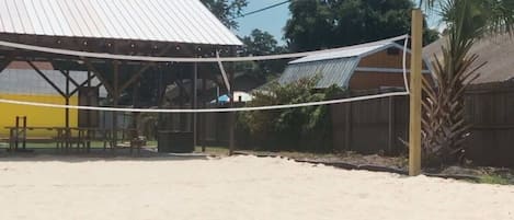 A sandy outdoor volleyball court with a metal roof structure, picnic tables, and a palm tree.