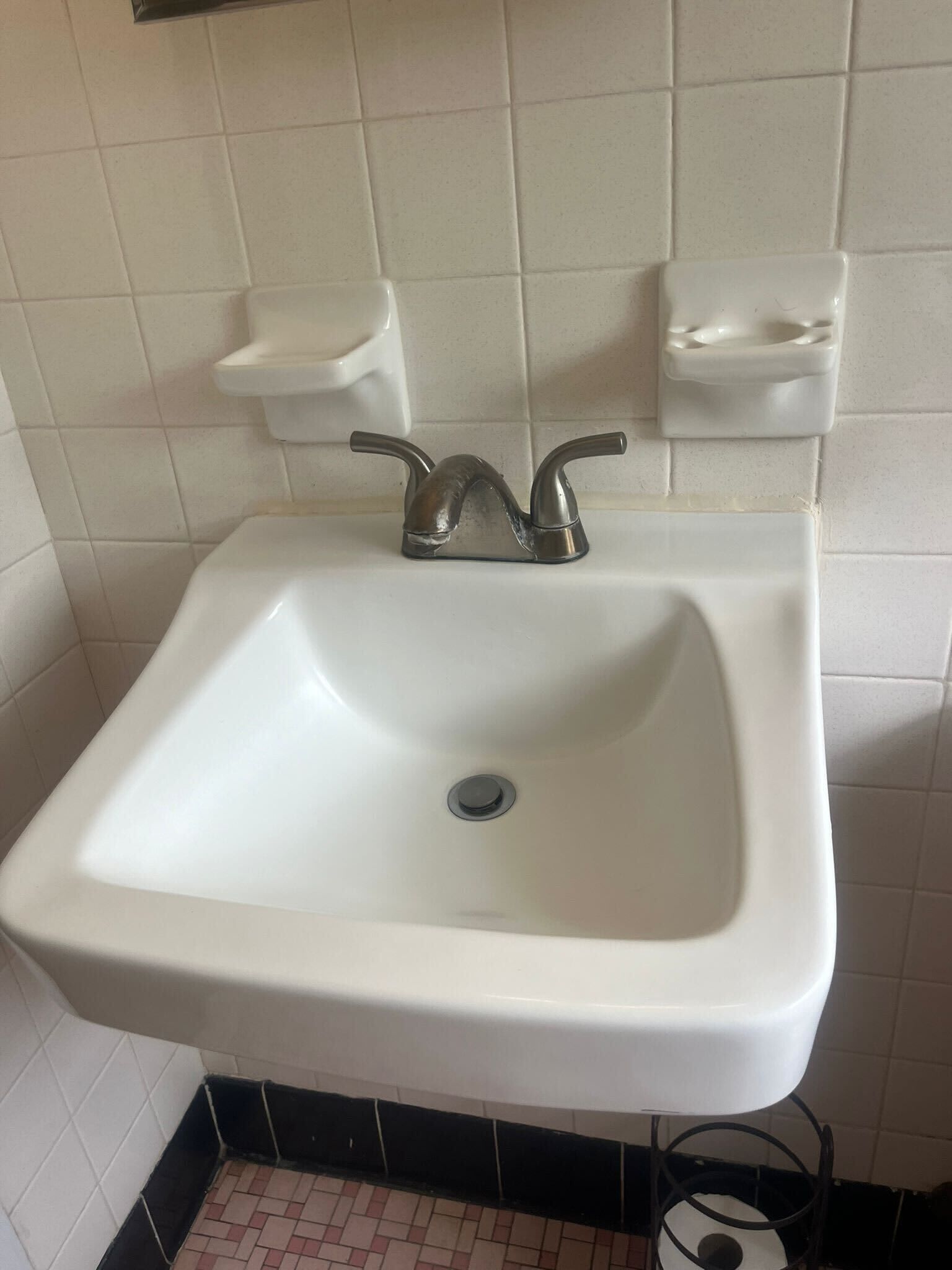 Ein weißes Waschbecken mit einem Wasserhahn, zwei Seifenspendern an der Wand und einem gefliesten Boden.