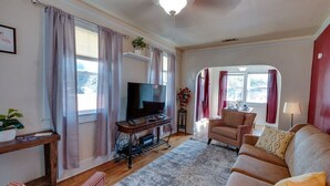 A living room with a television on a wooden stand, a sofa, a chair, and a small table with a lamp. - Dock&Volleyball! Near Downtown - Flamingo Flat (Savannah)