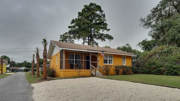 A yellow single-story building with a porch, a flag, and a gravel driveway.