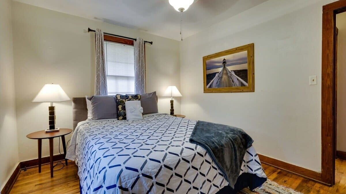 Un dormitorio con cama, dos lámparas de noche, una ventana con cortinas, un cuadro en la pared y suelo de madera.