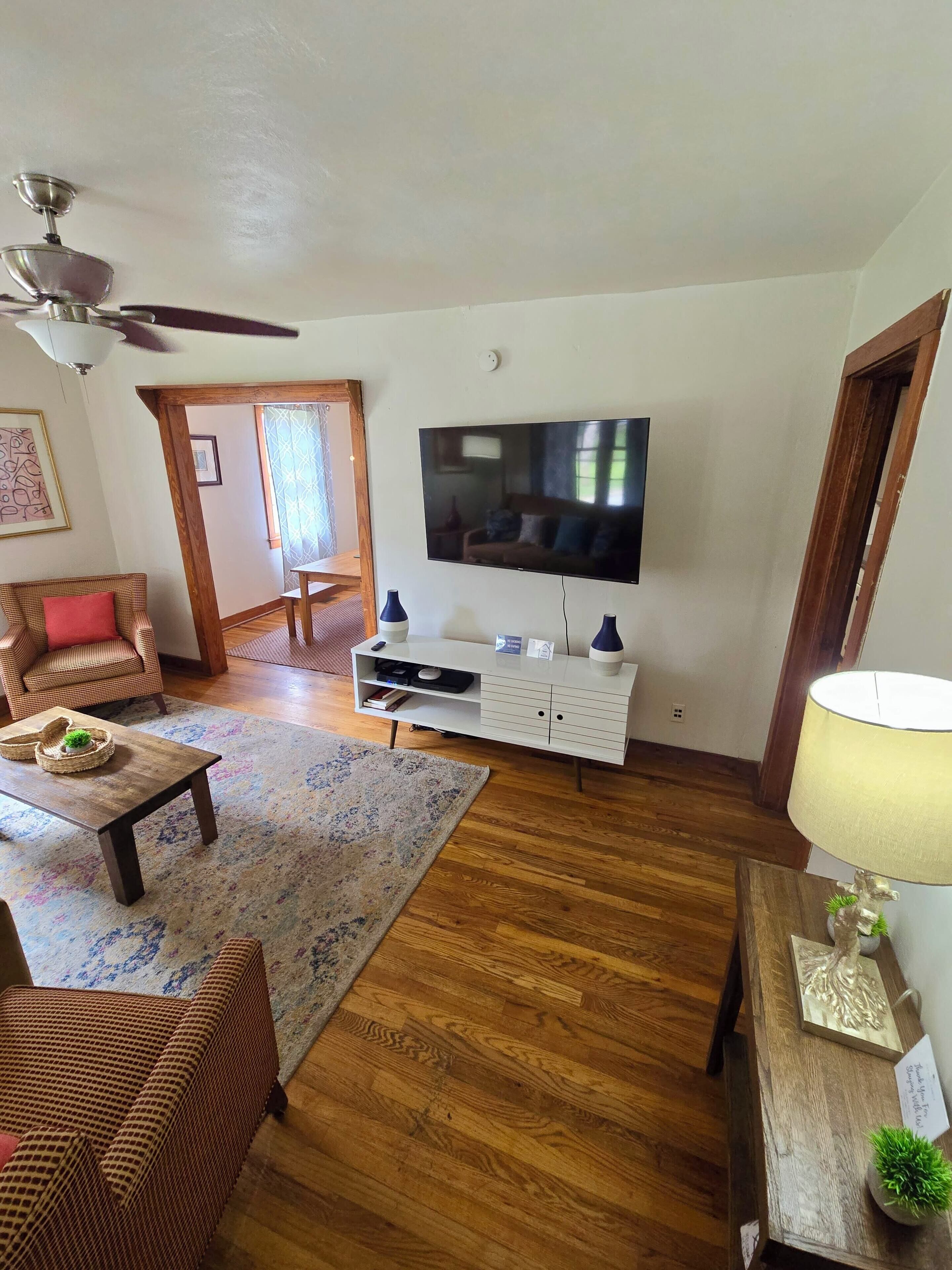 A living room with a television, wooden flooring, a ceiling fan, a sofa, a coffee table, and a lamp.