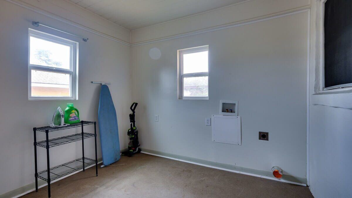 A room with two windows, a metal shelf with cleaning supplies, a vacuum cleaner, and a small electrical outlet on the wall.