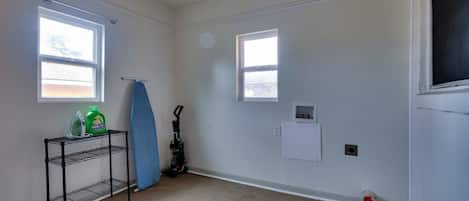 A room with two windows, a metal shelf with cleaning supplies, a vacuum cleaner, and a small electrical outlet on the wall.