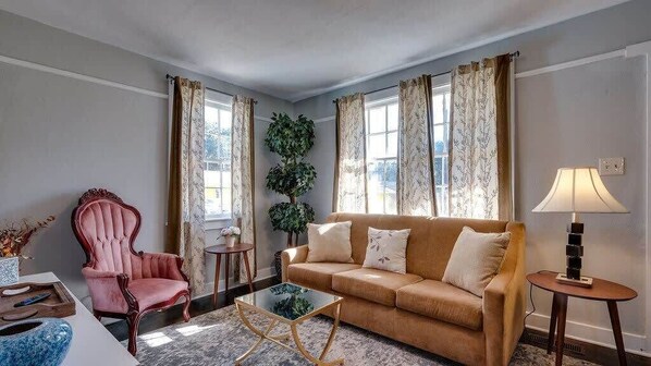 A living room with a pink chair, a beige sofa, a glass coffee table, and a lamp on a side table.