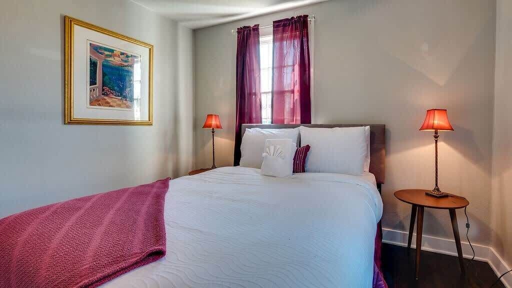 A neatly made bed with a white comforter and pillows, two bedside lamps, a framed picture on the wall, and a window with purple curtains.