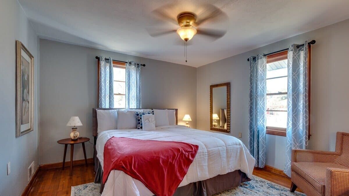 Ein Schlafzimmer mit einem Bett, einem Deckenventilator, einem Nachttisch mit Lampe, einem Sessel und einem Fenster mit Vorhängen.