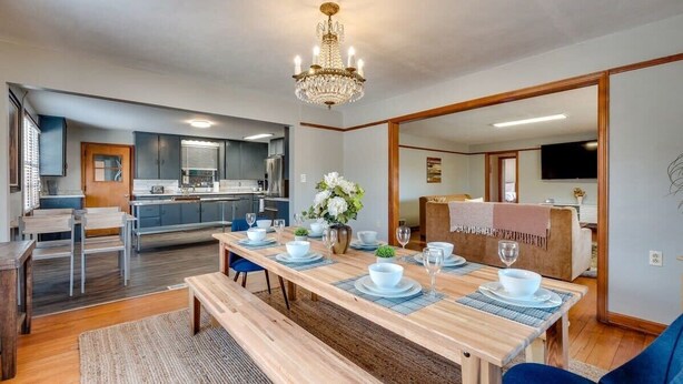 A modern dining area with a wooden table set for four, a chandelier, and a kitchen in the background. - Dock&Volleyball! Near Downtown - Totally Turquoise (Savannah)