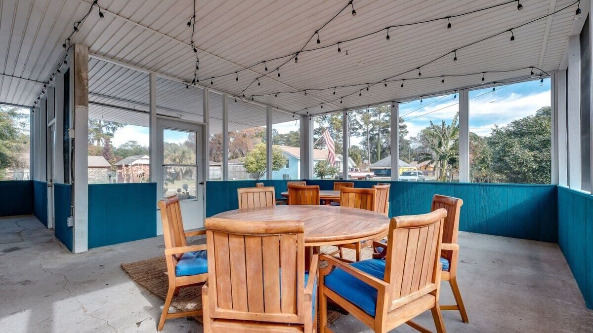 A covered outdoor seating area with a round wooden table and chairs, large windows, and a view of the surrounding area.