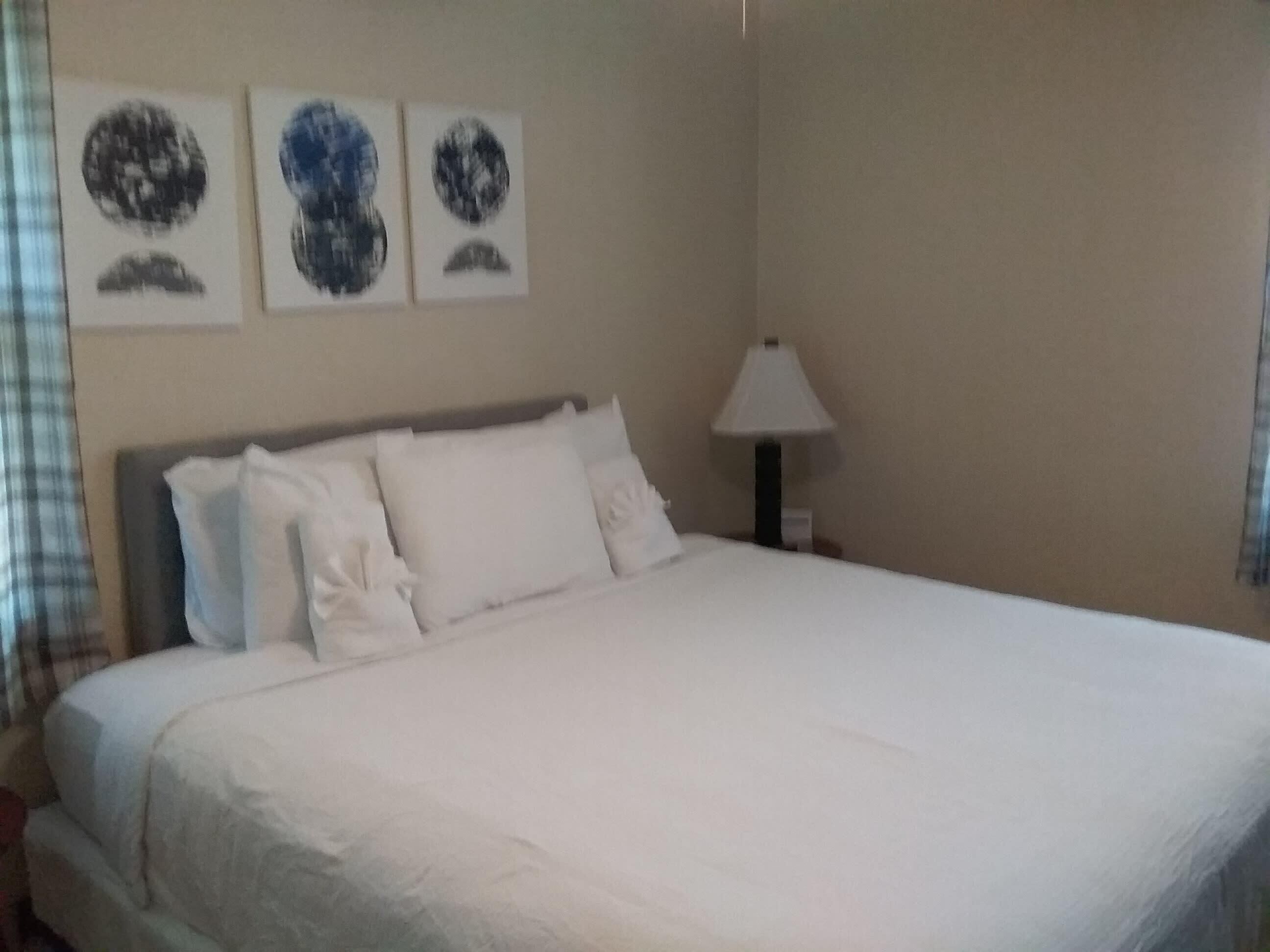 Un lit bien fait, avec du linge de lit blanc, dans une pièce au design simple et épuré. Trois œuvres d’art encadrées sont accrochées au mur, au-dessus du lit.