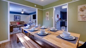 Un comedor con una larga mesa de madera dispuesta para seis personas, un sofá y una cocina al fondo.