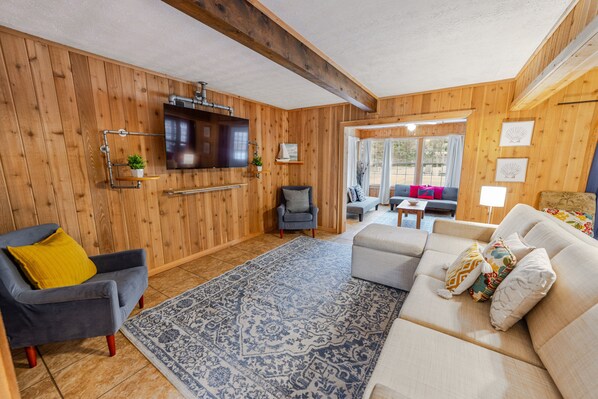 A cozy living room with wooden walls, a large sofa, a grey armchair, and a patterned rug.