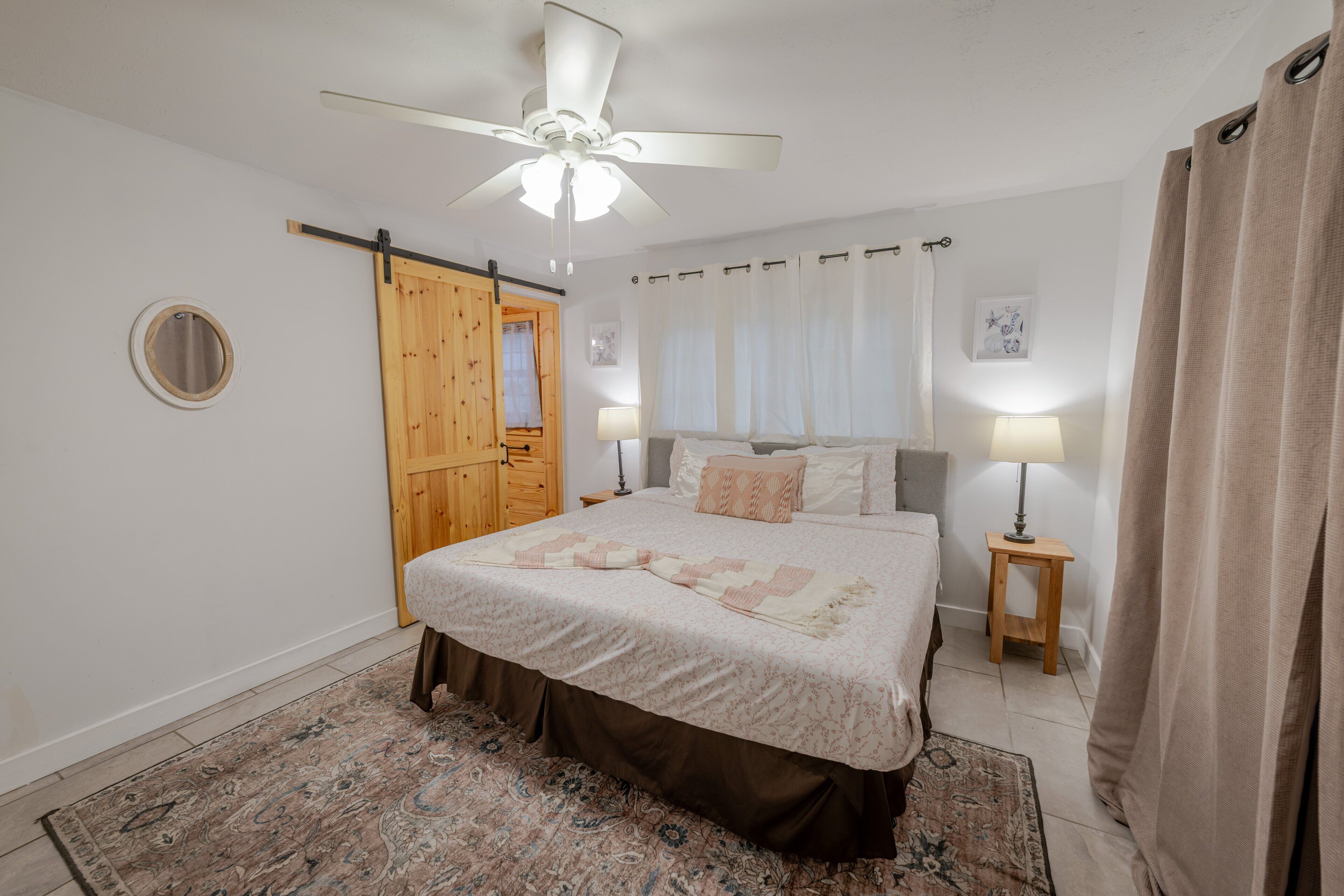 A bedroom with a bed, a ceiling fan, a wooden door, a small table with a lamp, and a rug.